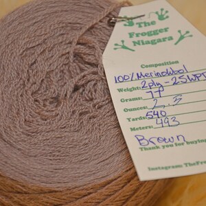 Może przedstawiać: Motka brązowej wełny merino 100%, 2-nitkowa, 25 WPI. Wełna jest nawinięta na kulkę i ma przyczepioną etykietę z informacjami: 77 gram&oacute;w, 2,5 uncji, 540 jard&oacute;w, 493 metr&oacute;w. Etykieta zawiera r&oacute;wnież napis "The Frogger Niagara" i "Dziękujemy za zakup recyklingu!"