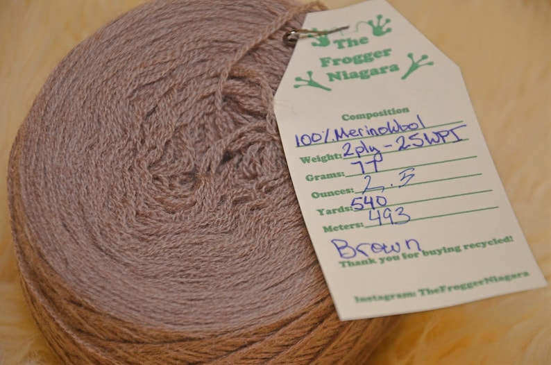 Może przedstawiać: Motka brązowej wełny merino 100%, 2-nitkowa, 25 WPI. Wełna jest nawinięta na kulkę i ma przyczepioną etykietę z informacjami: 77 gram&oacute;w, 2,5 uncji, 540 jard&oacute;w, 493 metr&oacute;w. Etykieta zawiera r&oacute;wnież napis "The Frogger Niagara" i "Dziękujemy za zakup recyklingu!"