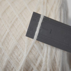 May include: A close-up of a white yarn ball with a ruler measuring one inch of the yarn. The ruler is marked in inches and 1/16ths of an inch.