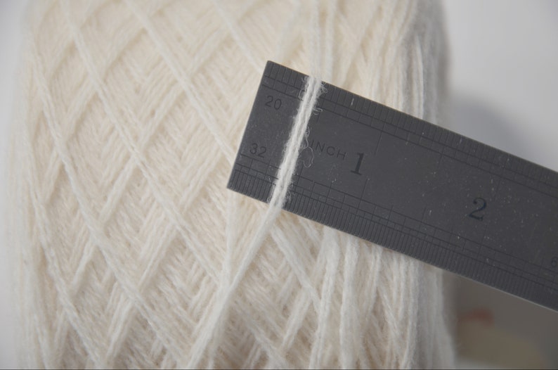 May include: A close-up of a white yarn ball with a ruler measuring one inch of the yarn. The ruler is marked in inches and 1/16ths of an inch.