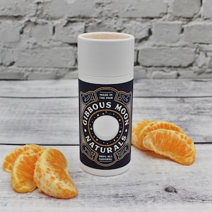 May include: A cylindrical container with a white top and a navy blue label featuring the text "Gibbous Moon Naturals" and "100% All Natural." The container is next to several orange tangerine segments.