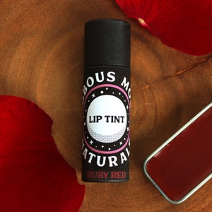 May include: A black tube of "Fabulous Moon" natural lip tint in "Ruby Red" with a white and pink label. A small, rectangular, red lip balm tin is next to the tube. Red rose petals are in the background.