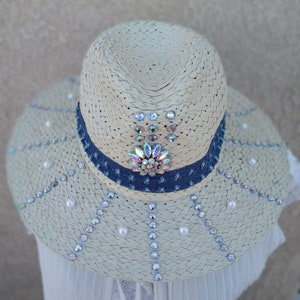 Op de afbeelding: Een witte strohoed met een blauwe denim band en parel- en regenboogkleurige strass steentjes.