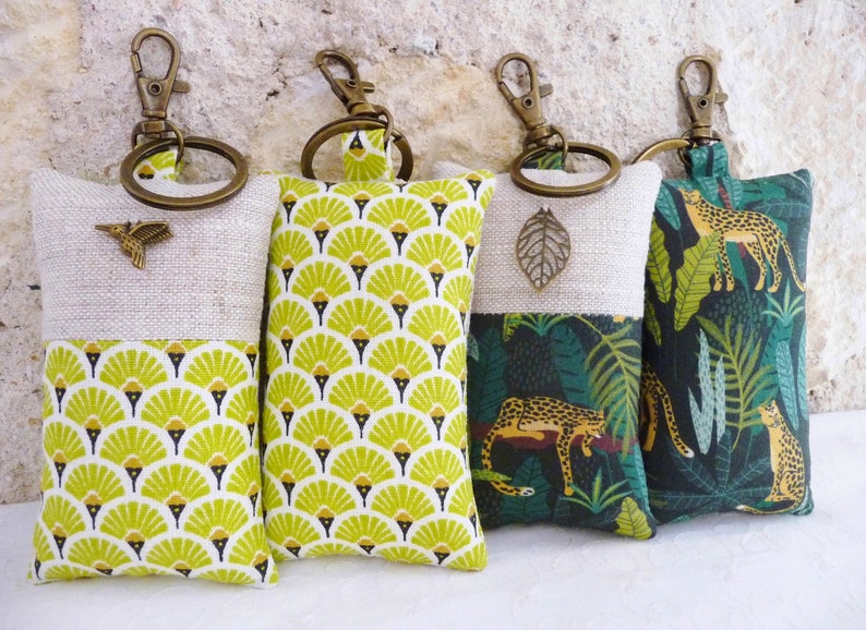 May include: Four keychains with different fabric designs. The keychains are made of linen and feature a variety of patterns, including a geometric design, a jungle scene, and a floral pattern. Each keychain has a brass ring and a metal charm.