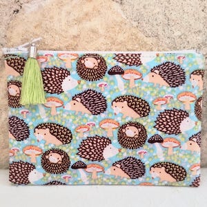 May include: A rectangular fabric zipper pouch featuring a repeating pattern of brown hedgehogs and orange mushrooms on a light blue background with green dots. A light green tassel is attached to the silver metal zipper pull at the top left.