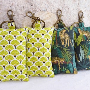 May include: Four keychains with different fabric patterns. Two keychains feature a green and yellow geometric pattern. The other two keychains feature a dark green background with a jungle pattern of leaves and leopards.