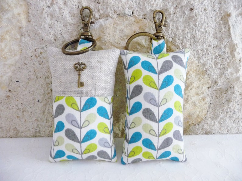 May include: Two keychains with fabric pouches. One pouch is beige linen with a vintage key charm and the other is white with a teal, green, and gray leaf pattern.