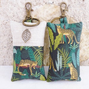 May include: Two keychains with a beige and green fabric design. One keychain features a leaf charm and the other features a repeating pattern of leopards and tropical leaves.