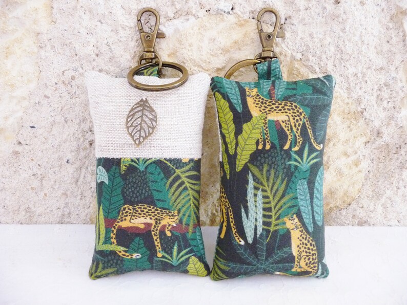 May include: Two keychains with a beige and green fabric design. One keychain features a leaf charm and the other features a repeating pattern of leopards and tropical leaves.