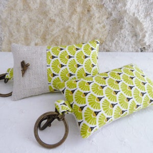 May include: Two keychains with small fabric pillows attached. The pillows are made of a light beige linen fabric with a green and black geometric pattern. One keychain has a small metal butterfly charm.
