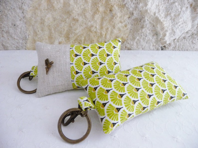 May include: Two keychains with small fabric pillows attached. The pillows are made of a light beige linen fabric with a green and black geometric pattern. One keychain has a small metal butterfly charm.