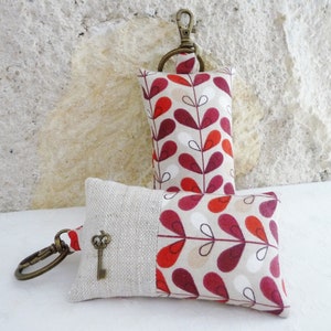 May include: Two keychains with a beige background and a red and white leaf pattern. The keychain on the left has a small key charm attached to it.