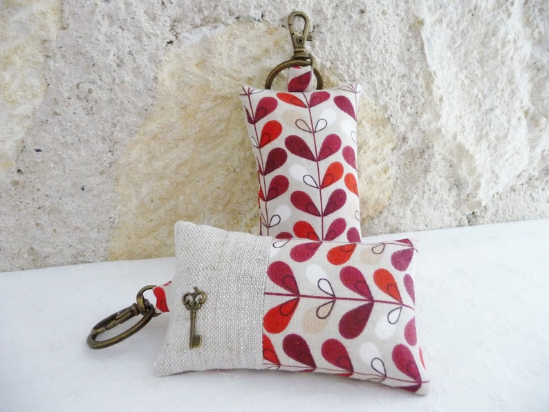 May include: Two keychains with a beige background and a red and white leaf pattern. The keychain on the left has a small key charm attached to it.