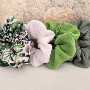 Peut inclure: Un ensemble de quatre chouchous en tissu disposés en ligne. Le premier présente un motif léopard vert et noir sur fond blanc. Le deuxième est blanc uni, le troisième est vert citron vif et le quatrième est d'un vert olive plus discret.