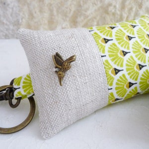 May include: A small, beige and green fabric key chain with a metal hummingbird charm. The fabric has a geometric pattern.