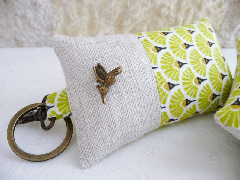 May include: A small, beige and green fabric key chain with a metal hummingbird charm. The fabric has a geometric pattern.
