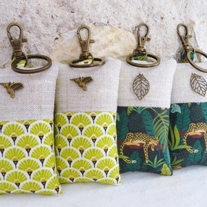May include: Four keychains with different designs. Each keychain is made of linen fabric with a different pattern on the bottom half. The top half is a natural linen color with a metal charm attached. The charms are a hummingbird, a leaf, a leopard, and a jungle scene.