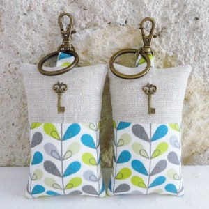 May include: Two keychains with a beige linen fabric pouch and a teal, green, and gray patterned fabric pouch. Each pouch has a small key charm attached to a brass ring.