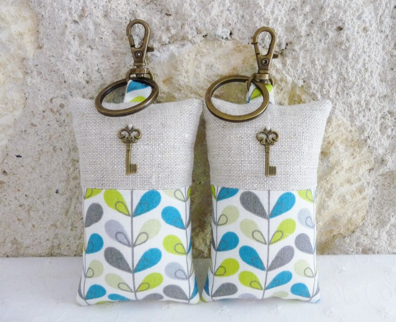 May include: Two keychains with a beige linen fabric pouch and a teal, green, and gray patterned fabric pouch. Each pouch has a small key charm attached to a brass ring.