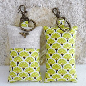 May include: Two keychains with a soft, padded design. One keychain is made of natural linen with a green and gold geometric pattern on the bottom half. The other keychain is made of a green and gold geometric fabric. Both keychains have a brass key ring.
