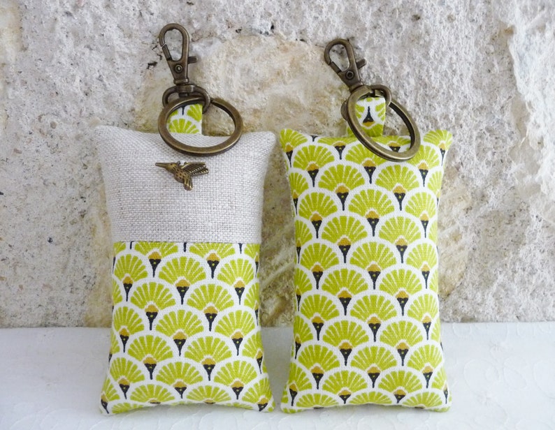 May include: Two keychains with a soft, padded design. One keychain is made of natural linen with a green and gold geometric pattern on the bottom half. The other keychain is made of a green and gold geometric fabric. Both keychains have a brass key ring.