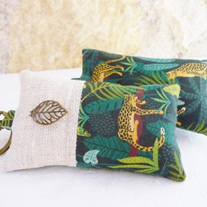 May include: A key chain with a small, beige fabric pouch. The pouch has a green and yellow fabric panel with a jungle print featuring leopards and leaves. A small, bronze leaf charm is attached to the pouch.