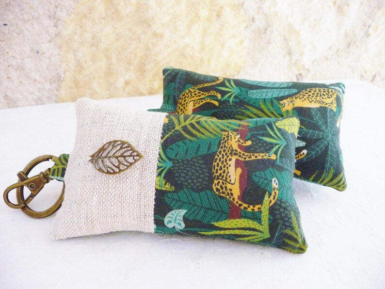 May include: A key chain with a small, beige fabric pouch. The pouch has a green and yellow fabric panel with a jungle print featuring leopards and leaves. A small, bronze leaf charm is attached to the pouch.