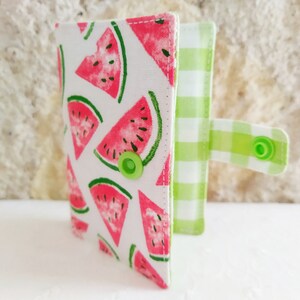 May include: A small, white fabric wallet with a watermelon print and a green and white checkered flap. The wallet has a green button closure.