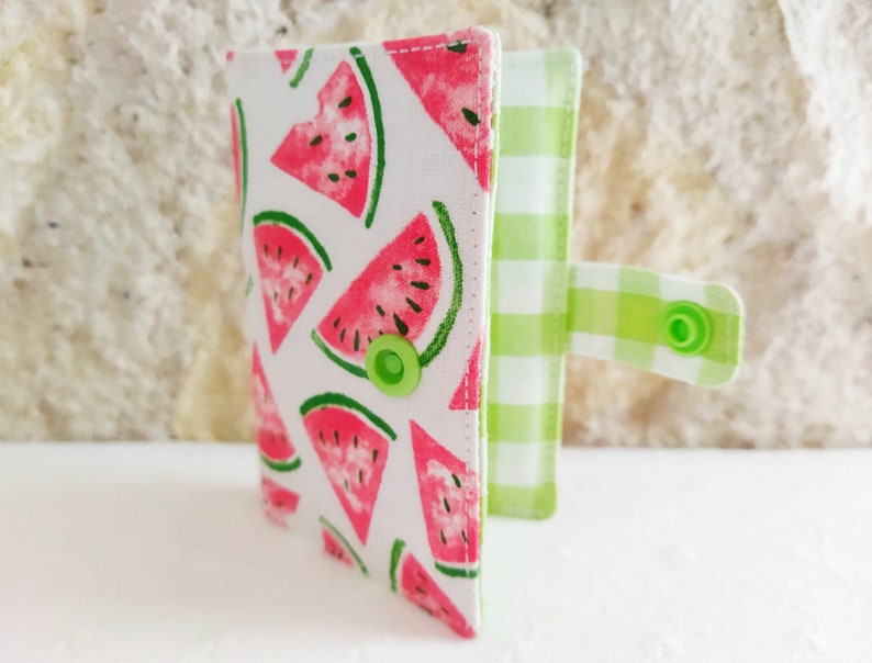 May include: A small, white fabric wallet with a watermelon print and a green and white checkered flap. The wallet has a green button closure.