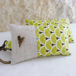 May include: A key chain with a small, brown bird charm attached to a beige and green fabric pouch. The pouch has a green and black geometric pattern on one side and a beige linen fabric on the other side.