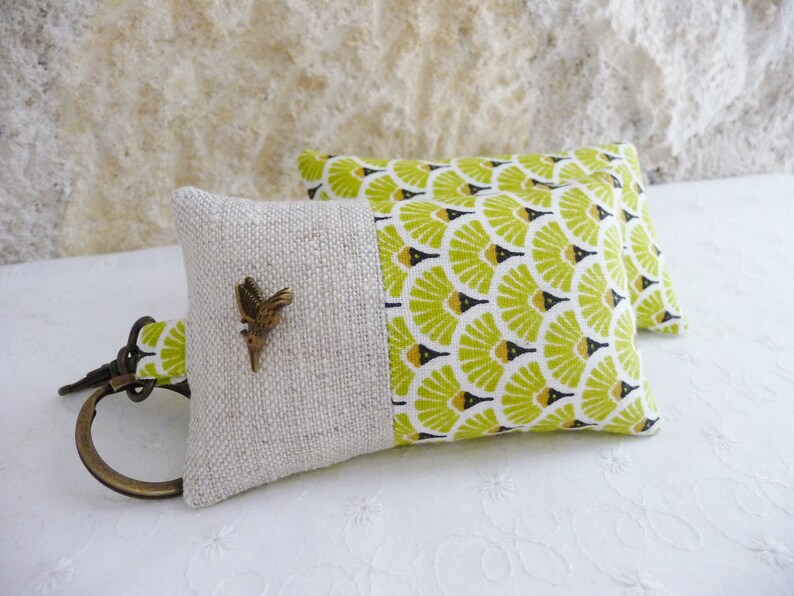 May include: A key chain with a small, brown bird charm attached to a beige and green fabric pouch. The pouch has a green and black geometric pattern on one side and a beige linen fabric on the other side.