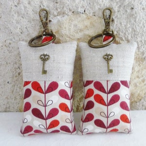 May include: Two keychains with a beige linen top and a red, white, and beige patterned bottom. Each keychain has a small antique-style key charm.