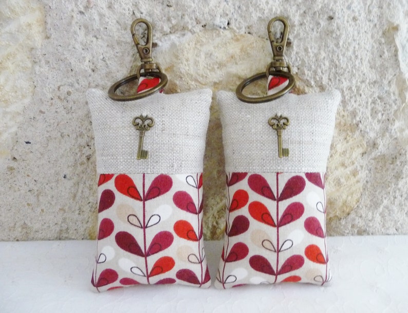 May include: Two keychains with a beige linen top and a red, white, and beige patterned bottom. Each keychain has a small antique-style key charm.