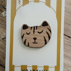 May include: Wooden cat face pin on a white card with gold stripes. The pin is light brown with dark brown stripes and closed eyes. The card has the text "LE PIN'S EN BOIS" and "KOADENN".