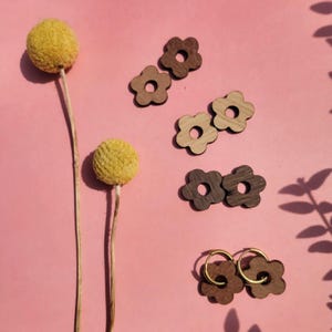Peut inclure: Une collection de breloques en bois en forme de fleur et une paire de boucles d'oreilles créoles avec des attaches en forme de fleur en bois. Les breloques sont de différentes nuances de brun avec une découpe circulaire centrale.