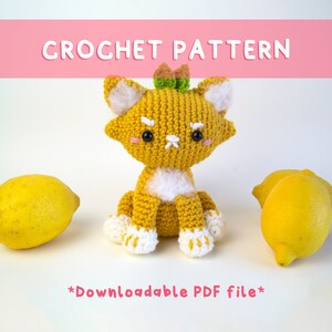 Op de afbeelding: Een gehaakte gele katachtige figuur met witte accenten, zittend tussen twee citroenen. De figuur heeft een groene en bruine kroon. De tekst "CROCHET PATTERN" staat boven de figuur en "*Downloadable PDF file*" eronder.