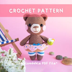 Könnte beinhalten: Ein gehäkelter Bär mit einem rosa Donut-Schwimmring und Badehose, der ein grünes Getränk hält. Der Bär hat einen rosa Hut und Rouge. Das Bild hat den Text "CROCHET PATTERN" und "*Downloadable PDF file*" auf rosa Hintergrund.
