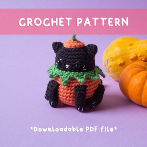 Puede incluir: Patrón de crochet para un gato negro vestido de calabaza. El gato está sentado sobre una superficie morada con dos calabazas en el fondo. El patrón es descargable como archivo PDF.