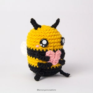 May include: A crocheted bee plush toy holding a pink heart. The bee is yellow and black with black eyes and a white wing.