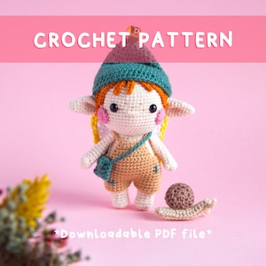 May include: A crocheted doll with orange hair, a teal hat, and a tan jumpsuit. The doll has large eyes, pink cheeks, and small wings. A small crocheted snail sits next to the doll. The words "CROCHET PATTERN" and "Downloadable PDF file" are displayed.
