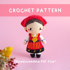 May include: A crocheted doll wearing a red headscarf and dress with yellow and black accents. The doll has a gold heart-shaped necklace and black shoes. The text "CROCHET PATTERN" is at the top of the image.