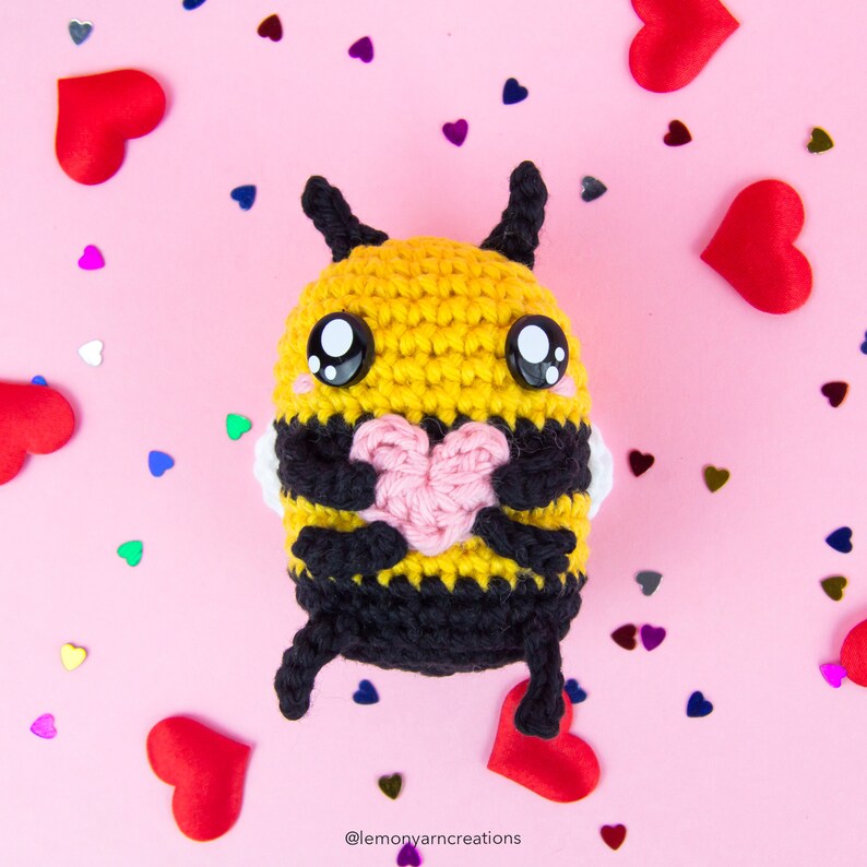 May include: A crocheted bee holding a pink heart in its arms. The bee is yellow and black with big black eyes. The bee is surrounded by small red hearts on a pink background.