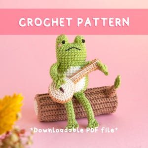 Op de afbeelding: Een haakpatroon met een groene kikker met een witte buik die op een bruine boomstam zit en een beige banjo bespeelt. De tekst CROCHET PATTERN staat bovenaan en *Downloadable PDF file* staat onderaan op een roze achtergrond.