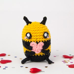 May include: A crocheted bee holding a pink heart. The bee is yellow and black with large black eyes. The bee is surrounded by small hearts.
