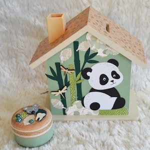 Op de afbeelding: Een houten spaarpot in de vorm van een huis met illustraties van een panda, bamboe en libellen op een lichtgroene achtergrond, vergezeld van een klein rond houten bakje versierd met een muisfiguurtje, blauwe kralen en een witte tandvormige uitsparing.