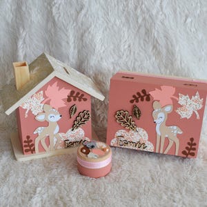 May include: A wooden house-shaped piggy bank and a rectangular wooden box, both painted dusty pink and decorated with paper deer, floral leaves, and the name Esm&eacute;e. A small round pink container with a mouse figure sits in the foreground.