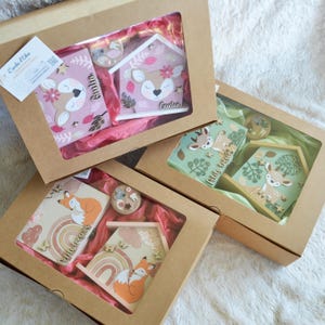 May include: Three brown cardboard gift boxes with clear windows, each containing a wooden house-shaped box and a small round container. The sets feature illustrations of deer and foxes in pink, green, and beige, with wooden accents and text.