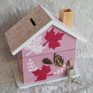 May include: A pink wooden house-shaped piggy bank with a floral patterned roof, a wooden chimney, and a coin slot. The exterior is decorated with glittery red and light pink leaf cutouts, and it features a metal padlock with a key attached.