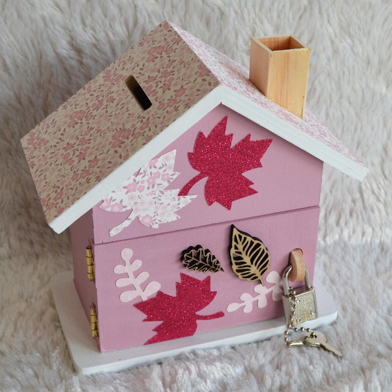 May include: A pink wooden house-shaped piggy bank with a floral patterned roof, a wooden chimney, and a coin slot. The exterior is decorated with glittery red and light pink leaf cutouts, and it features a metal padlock with a key attached.