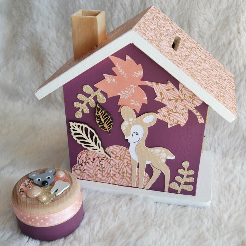 May include: A wooden house-shaped piggy bank painted deep purple with a light pink patterned roof, featuring a cutout deer, gold glitter leaves, and a chimney. Beside it sits a small round wooden container with a mouse and tooth embellishment.